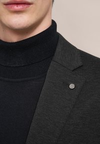 Schwarzer Blazer aus einem strukturierten Stoff, kombiniert mit einem enganliegenden schwarzen Rollkragenpullover. Der Blazer hat einen silbernen Akzentknopf am Revers.