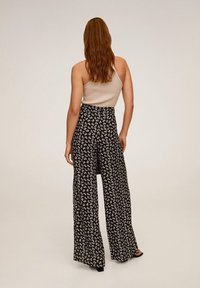 Kobieta z długimi rudymi włosami stoi tyłem, ubrana w kremowy top na jedno ramię i czarne szerokie spodnie w biały kwiatowy wzór.