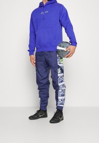 Felpa blu con tasca frontale, pantaloni navy con loghi grafici "SPURS", e sneakers nere. Tiene in mano una palla grigia con texture.