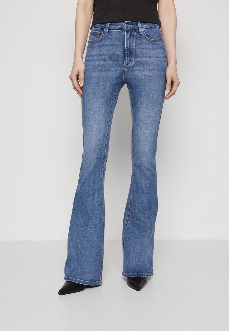 Personne portant un jean bleu évasé et des chaussures noires pointues, debout devant un fond blanc uni, visible des épaules aux pieds.