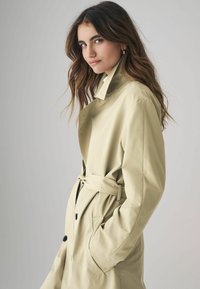 Kerge beež trench coat, millel on vöökoht, suured taskud ja klassikaline krae. Valmistatud sujuvast tekstureeritud kangast.