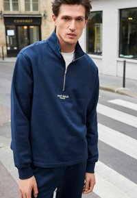 Felpa zippata blu navy con colletto alto, caratterizzata da un piccolo ricamo "SAINT-MALO FRANCE". Tessuto morbido e testurizzato, con polsini a coste.