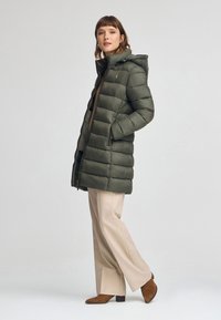 Olivgrön puffjacka med huva, med quiltade sektioner och sidofickor. Buren med beige vida byxor och bruna klackstövlar.