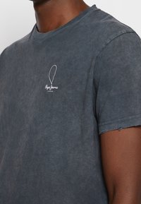 Šedé tričko s kulatým výstřihem vyrobené z měkkého materiálu, se zdrsněným lemem a malým bílým logem na levé části hrudi.