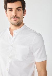 Rövid ujjú, fehér pamut ing, álló gallérral, elöl gombos záródással, és egy kis zsebbel a bal oldalon. Sima textúra.