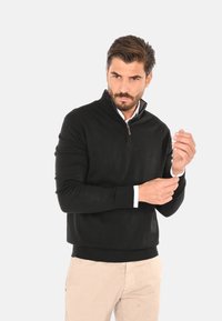Maglione zip-up nero con colletto alto, realizzato in materiale morbido, dotato di polsini e orlo a costine, indossato sopra una camicia bianca con colletto.