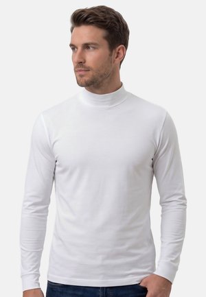 Mann trägt ein weißes, langärmeliges Rollkragenhemd und blaue Jeans, schaut nach links vor einem einfarbigen hellen Hintergrund.