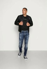 Jovem vestido com uma jaqueta de couro preta, uma t-shirt preta estampada, jeans skinny azuis desgastados e sapatilhas pretas, encostado a uma parede branca.