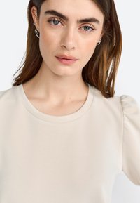 Haut beige avec un col rond et des manches bouffantes, fabriqué en tissu doux. Présente une texture lisse et un design minimaliste.