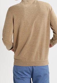 Camicia beige a maniche lunghe con finitura testurizzata, dotata di colletto girocollo e polsini e orlo a costine, abbinata a pantaloni corti azzurri chiari.
