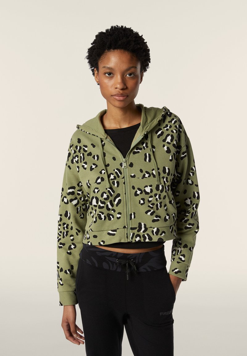 Hoodie vert raccourci avec imprimé léopard noir et blanc, fermeture éclair à l'avant et capuche avec cordon de serrage. Porté avec un pantalon noir à taille avec motif.