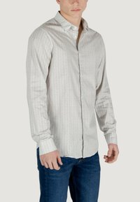 Camicetta chiara con bottoni, caratterizzata da un sottile motivo geometrico, maniche lunghe e colletto aperto, abbinata a jeans blu.