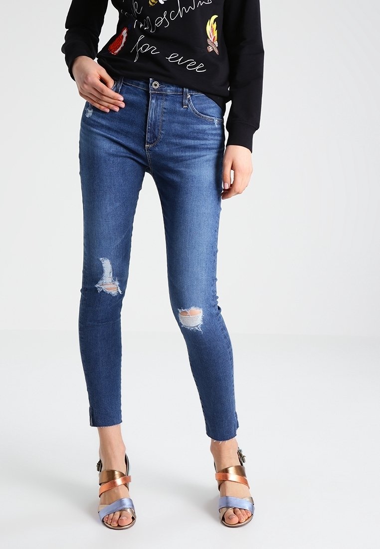 Jeans de mezclilla azul ajustados con detalles desgastados en las rodillas, combinados con un sudadero negro y sandalias de tiras con acentos metálicos.