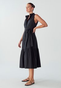 Abito senza maniche di colore nero con colletto, apertura frontale con bottoni, vita con cintura e gonna a strati. Tessuto liscio con una leggera lucentezza e texture minima.