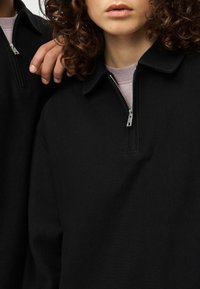 Μαύρο ριμπ πουλόβερ με φερμουάρ στον γιακά. Διαθέτει λεία υφή και εφαρμοστή σιλουέτα. Συνδυάζεται με ανοιχτόχρωμο τοπ από κάτω.