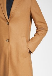 Camelkleurige jas van zachte stof, met een gesneden revers, twee knopen en een zijzak voor praktisch gebruik.