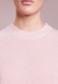 Jasnoróżowy sweter z prążkowanej dzianiny o szerokim, zaokrąglonym dekolcie i miękkiej fakturze. Posiada pionowy wzór prążków i minimalne detale.