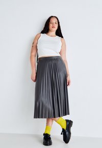 Gonna midi grigia plissettata realizzata in materiale lucido, abbinata a una blusa bianca senza maniche e scarpe nere. Calzini gialli aggiungono un contrasto di colore.