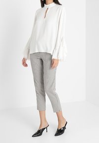Blusa bianca con collo alto e dettaglio a occhiello, abbinata a pantaloni a scacchi bianchi e neri. Mules neri lucidi completano il look.