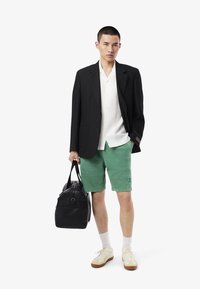 Blazer noir, chemise blanche à boutons, shorts verts, portant un sac noir. Chaussettes blanches avec des baskets beiges. Lignes épurées et coupe décontractée.