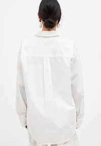 White long-sleeve shirt with a relaxed fit, back pleat detail, and buttoned cuffs. Smooth cotton fabric with a classic collar design.