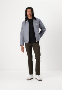 Grå zip-up jakke lavet af robust stof, sort skjorte under, grønne bukser, og hvide sneakers med grå detaljer. Stående stilling.
