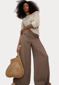 Un pull en maille beige clair avec des manches retroussées, associé à un pantalon en lin marron ample. Un sac en toile beige tenu dans la main droite.