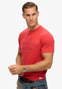Red cotton t-shirt with short sleeves, featuring a dark print that reads "SUPERDRY & CO" and "EST 1954." Worn with a blue plaid shirt tied around the waist.