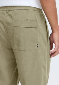 Beige Cargo-Hosen verfügen über eine Rückentasche mit Klappe, einen elastischen Bund und ein kleines schwarzes Logotag an der Seite. Glatte Stofftextur.
