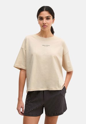 Mujer de cabello oscuro con una camiseta beige de Marc O’Polo y pantalones cortos negros con patrones de diamantes blancos, de pie con una mano en el bolsillo.