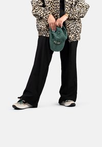 Veste bomber en imprimé léopard, pantalon noir à jambe large, casquette verte tenue à la main et baskets multicolores avec des lacets contrastants.