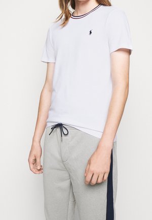 Person trägt ein weißes kurzärmeliges T-Shirt mit marineblauem Besatz und Logo, kombiniert mit hellgrauen Jogginghosen mit einem dunkelblauen Seitenstreifen und Kordelzug.