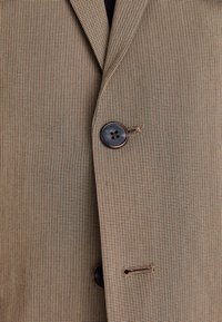Brauner Blazer mit Hahnentrap Muster aus strukturiertem Stoff, ausgestattet mit einem einzelnen braunen Knopf und einem detaillierten Stich am Revers.