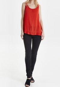 Top rosso senza maniche con orlo a balze, abbinato a pantaloni neri slim-fit e sandali neri con tacco aperto; texture del tessuto liscia.