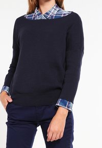 Granatowy sweter z prążkowanego materiału z otwartym dekoltem, nałożony na niebiesko-białą koszulę w kratę. Posiada długie rękawy i luźny krój.