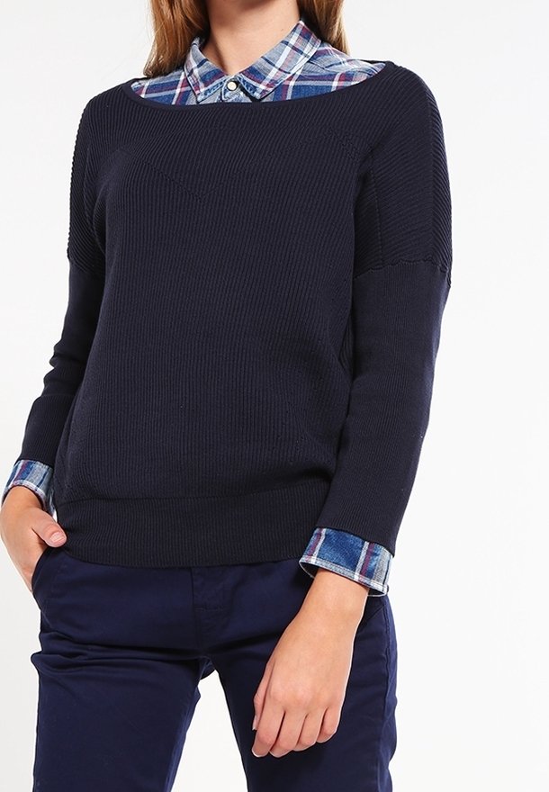 Granatowy sweter z prążkowanego materiału z otwartym dekoltem, nałożony na niebiesko-białą koszulę w kratę. Posiada długie rękawy i luźny krój.
