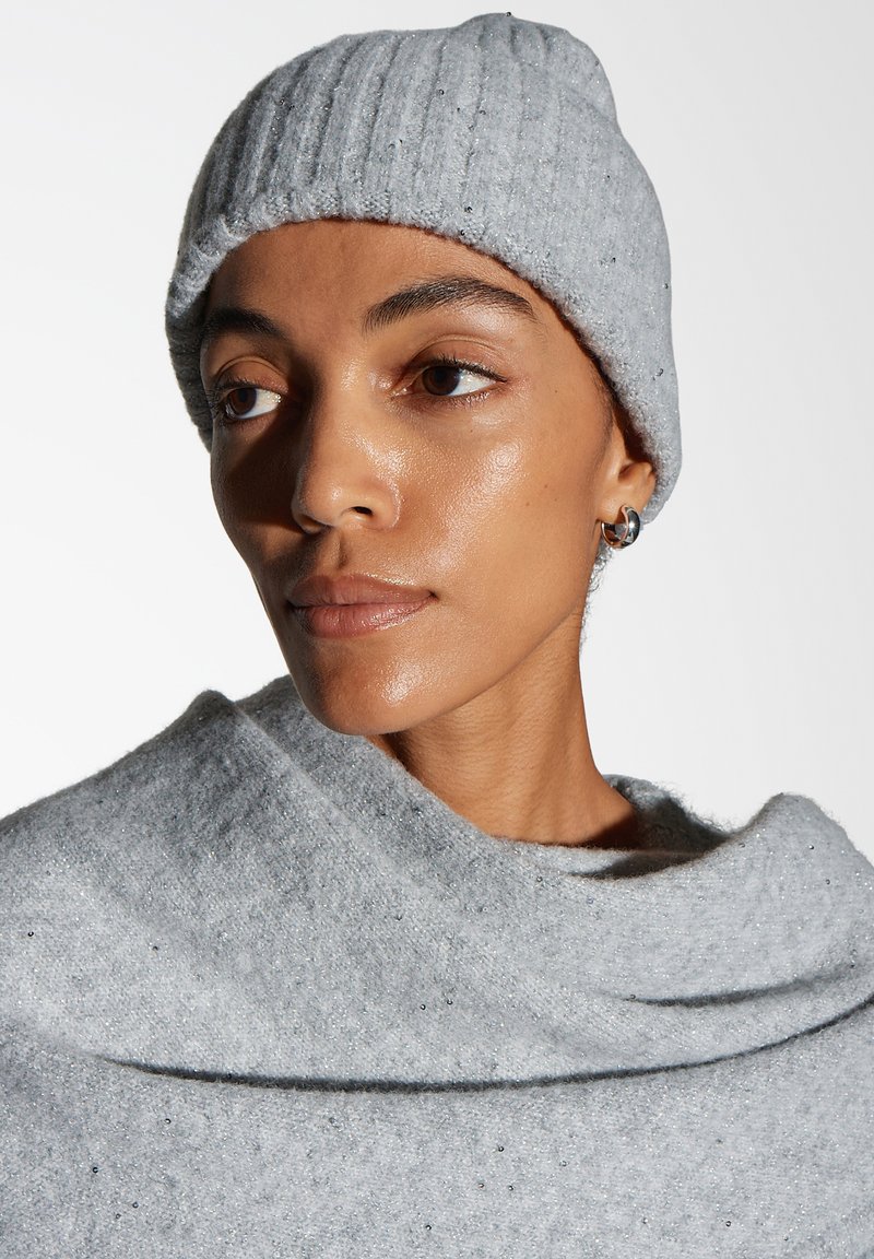 Frau, die eine hellgraue Strickmütze und einen passenden Pullover mit kleinen glänzenden Verzierungen trägt und leicht zur Seite schaut.