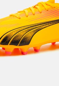 Botín de fútbol amarillo brillante con detalles en negro y tacos de suela rosa. Cuenta con una superficie texturizada y cordones amarillos a juego.