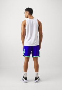 Top sin mangas blanco y pantalones cortos azules con detalles verdes, con textura de malla. Calcetines deportivos blancos y zapatos negros con detalles blancos.