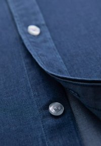 Una camicia in denim blu scuro in primo piano, con una texture liscia, colletto a punta e due bottoni argentati con dettagli incisi.