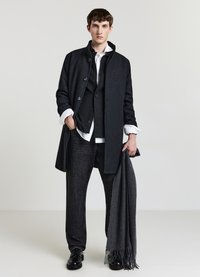 Cappotto di lana nero con collo alto, chiusura con bottoni, indossato sopra un gilet scuro e una camicia bianca. Jeans scuri e sciarpa grigia con frange.