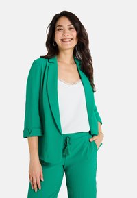 Blazer verde con mangas arremangadas y una camiseta sin mangas blanca con encaje, combinada con pantalones verdes a juego que tienen un cordón con borlas.