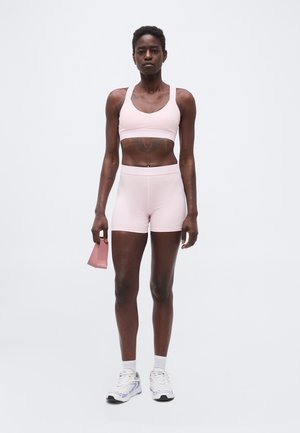 Kobieta w jasnoróżowym sportowym biustonoszu i szortach trzymająca różową taśmę oporową, stojąca na tle jednolitego białego tła.