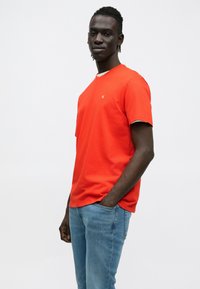 Raudonas medvilninis marškinėlis su trumpomis rankovėmis, apvalia iškirpte ir mažu logotipu krūtinės srityje. Derinamas su šviesiai mėlynomis džinsinėmis kelnėmis.