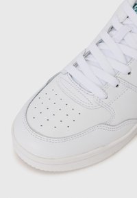 White sneaker with smooth leather material, featuring perforated toe box, flat laces, and rounded rubber sole. Stitching visible along edges.