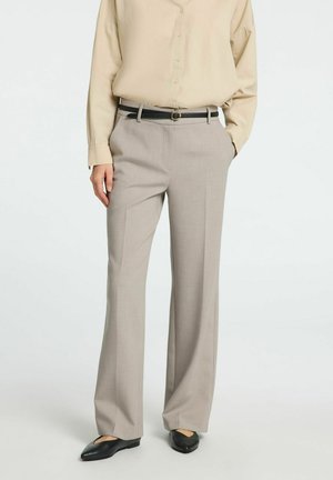 Personne portant un pantalon large beige clair avec ceinture noire, chemise crème boutonnée rentrée, et chaussures plates noires, debout sur fond blanc.