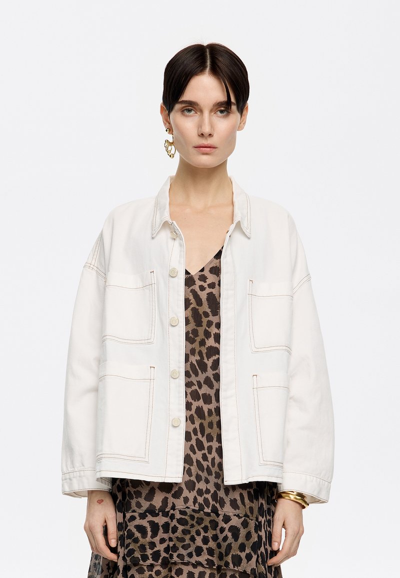 Veste en denim blanc avec quatre poches plaquées, fermeture à boutons et détails de couture visibles, portée sur une robe à imprimé léopard.