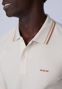 Polo shirt crème en tissu texturé, avec un col à rayures marron et beige et trois petits ours brodés marron.