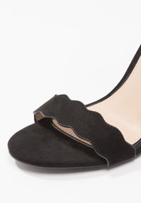 Sandale à talon haut en suède noir avec un design de bretelles à bord festonné et une semelle intérieure nude. Texture lisse avec une forme ouverte et élégante.