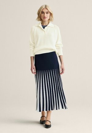Lyshåret kvinde iført en hvid ribstrikket sweater og et sort-hvidt plisseret midinederdel med sorte sandaler med remme, stående mod en cremefarvet baggrund.